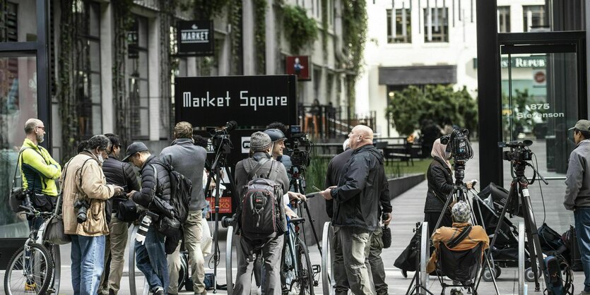 Journalist*innen mit Kameras vor Gebäuden, dazu das Schild Market Square