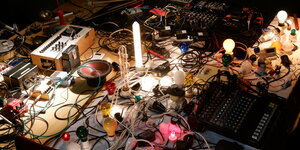 Analoges Equipment für elektronische Musik liegt auf einem Tisch. Es sind meherer Mischpulte und viele bunte Kabel zu sehen. Dazwischen stehen leuchtende Glühbirnen