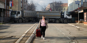 Eine Frau im rosa Pullover geht über eine leere Straße, im Hintergrund blockieren 2 LKWs die Straße