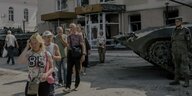 Menschen stehen in einer Warteschlange, hinter ihnen ein zerstörtes Haus, am Bildrand ein Panzer