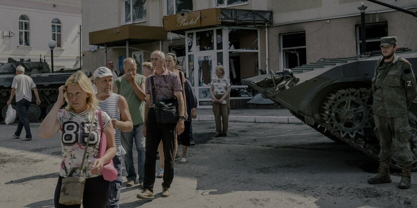 Menschen stehen in einer Warteschlange, hinter ihnen ein zerstörtes Haus, am Bildrand ein Panzer