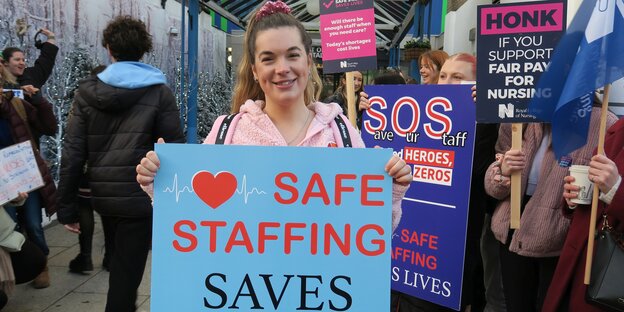 Laura Tossel hält lächelnd ein Schild ind die Kamera mit der englischen Aufschrifft: Sicheres Personal rettet Leben