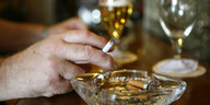 Hand mit Zigarette über Aschenbecher, im Hintergrund Biergläser