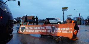Drei Klimaaktivist*innen sitzen auf einer Straße, vor sich haben sie ein organgenes Transparent mit der Aufschrift: "Letzte Generation vor den Kipppunkten"