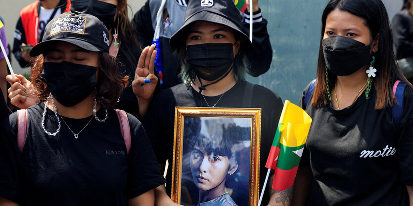 Drei Demonstrantinen mit medizinischen Masken, eine hält ein Bild Aung San Suu Kyis hoch, das sie in jungen Jahren zeigt.