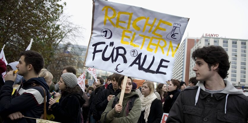 Eine Frau trägt bei einer Demonstration ein Schild mit der Aufschrift "Reiche Eltern für alle"