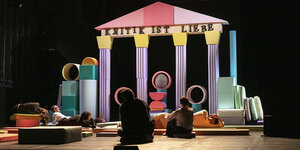 Ein bunter Tempel aus Bausteinen auf einer Bühne mit dem Schriftzug "Kritik ist Liebe"