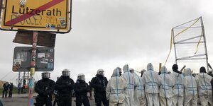 Polizei und Menschen in weißen Overalls stehen sich gegenüber. Ein Ortsschild zeigt "Lützerath" durchgestrichen, drüber estht geschrieben: "Bulle"