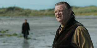 An der Küste am Strand sieht man einen Mann, Brendan Gleeson, im Vordergrund, weiter hinter nähert sich ein zweiter