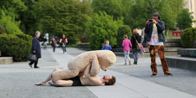 In einer begrünten Fussgängerzone liegt ein Tänzer auf dem Boden, über ihm ein lebensgroßer Teddy