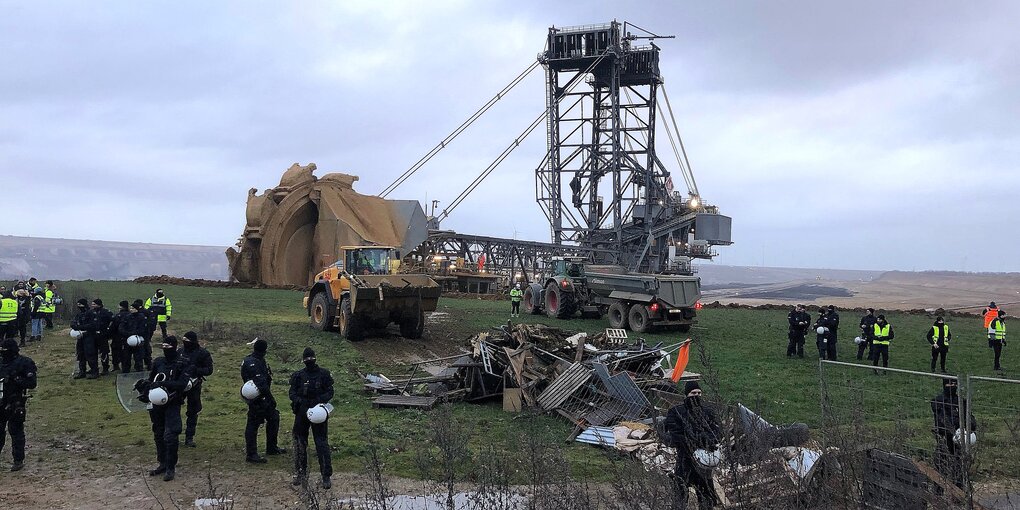 Kohlebagger im Hintergrund auf einem Feld, davor Polizei, Zäune und Sperrmüll