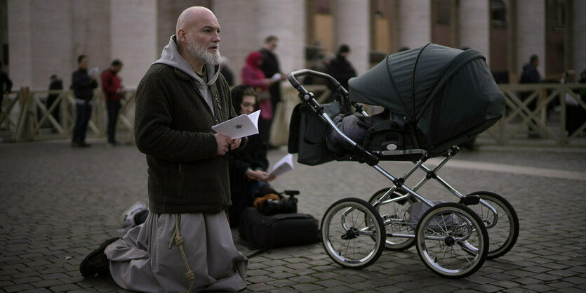 Ein Mann in grauer Kutte kniet auf dem Boden und betet, neben ihm eine Frau und vor ihm ein Kinderwagen