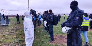 Demonstrant*innen in weißen Schutzanzügen stehen schwarz vermummten Polizeibeamten auf einem freien Feld gegenüber