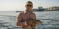 Daniel Craig als Benoit Blanc in „Glass Onion“. Er trägt ein rosa Hemd und ein Tuch um den Hals. Im Hintergrund: Wasser und ein entferntes Ufer.