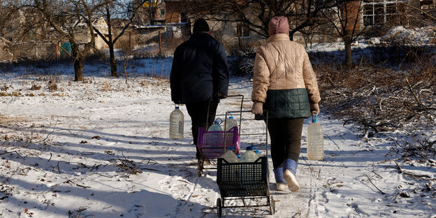 2 Personen gehen mit Einkaufswagen beladen mit Wasserflaschen durch den Schnee