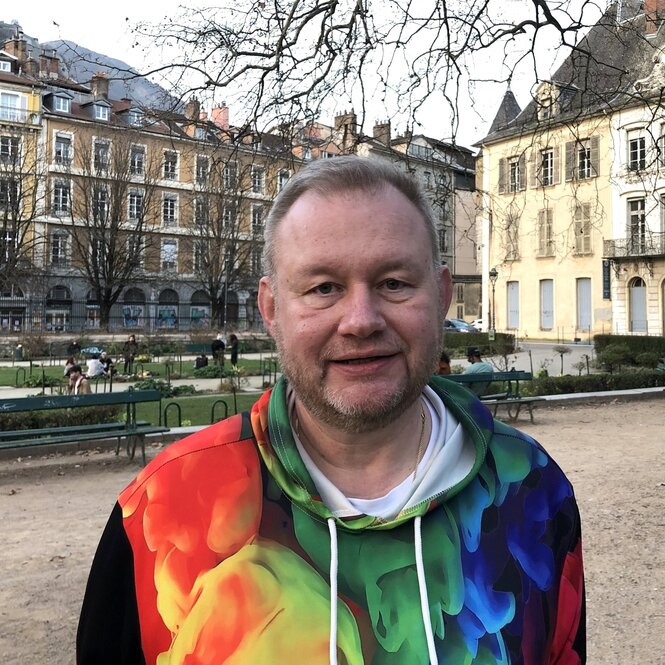 Holger Allmenroeder steht in einem bunten Shirt vor einem Gebäudeensemble