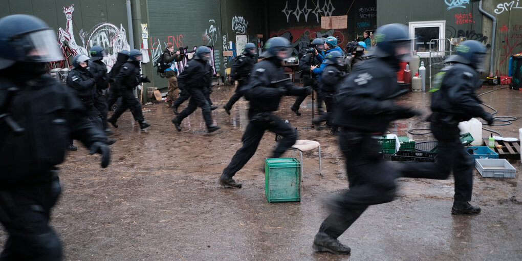 Lützerath: Mindestens ein Dutzend Polizisten, die in schwarzen Einsatzklamotten und mit Helm von links nach rechts über matschigen Boden rennen. Dazwischen ein paar wenige Menschen, die die Szene fotografieren oder filmen, und ein paar herumliegende Kisten. Im Hintergrund: grau-grüne Wände auf denen Graffitis sind.