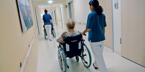 Zwei Mitarbeiterinnen schieben zwei Patientinnen in Rollstühlen durch einen Krankenhausflur.
