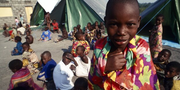 Ruandische Flüchtlingskinder. Eines steht im Vordergrund und schaut in die Kamera. Weitere, vor Zelten sitzende Kinder im Hintergrund.