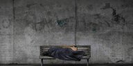 Ein Mensch liegt, in eine Decke gewickelt, auf einer Bank. Im Hintergrund eine graue Mauer
