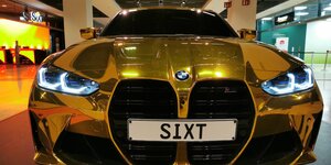 Ein goldener BMW ist in einer Flughafenhalle zu sehen. Auf dem Nummernschild steht: SIXT
