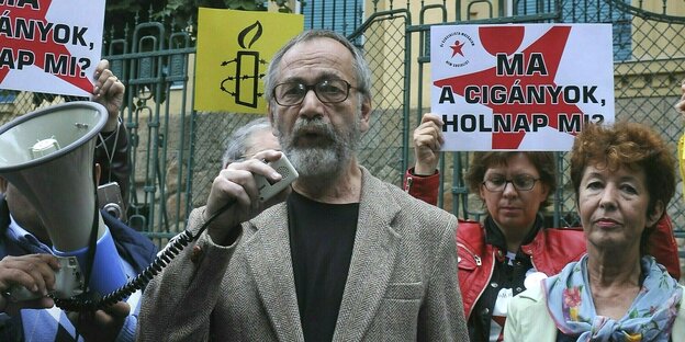 Gaspar Miklos Tamas spricht in ein Megafon umringt von Menschen, die Schilder hochhalten