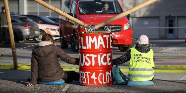 Zwei Personen sitzten auf dem Boden. In ihrer Mitte steht eine rote Tonne, auf der in weißer Schrift Climate Justice geschrieben steht.
