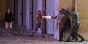Ein Polizist schießt während Demonstranten Steine werfen