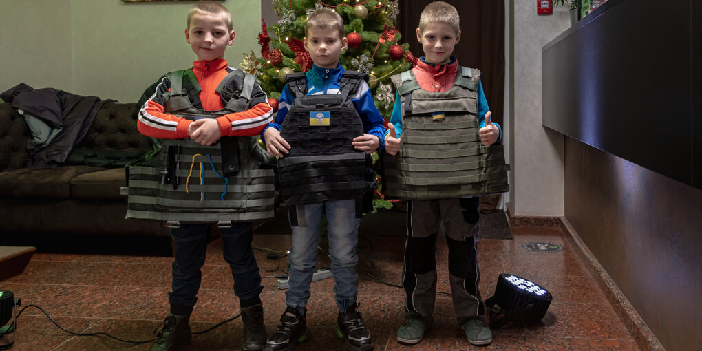 3 kleine Jungen in Schutzwesten stehen breitbeinig vor einem Weihnachtsbaum