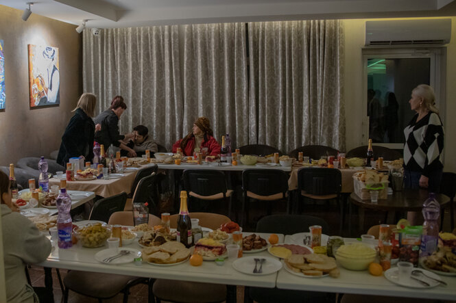 In einem Speisesaal ist ein Tisch festlich mit Essen und Sekt eingedeckt, traurige Menschen im trüben Licht im Raum