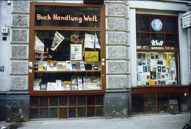 Ladenfront von Buch Handlung Welt, im Schaufenster sind Bücher zu sehen, ins Schaufenster ist eine taz.die tageszeitung gehängt, darum steht in Hand geschrieben "Jetzt täglich". Daneben steht "Neu! Fotokopie -, 50"