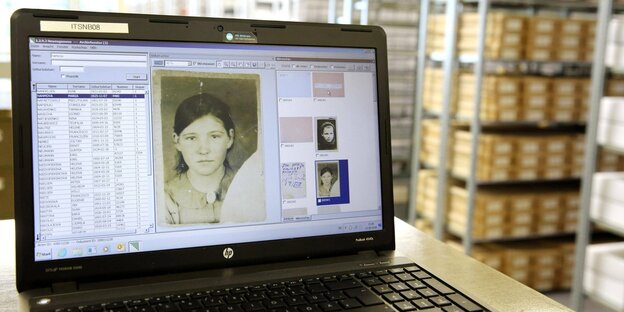 Auf einem Laptopbildschirm ist das Bild einer Frau zu sehen. Im Hintergrund stehen Regale mit Archivdokumenten