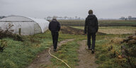 Zwei menschen gehen über ein Feld, links ist ein mit einer Plastikplane verkleidetes Gewächshaus zu sehen