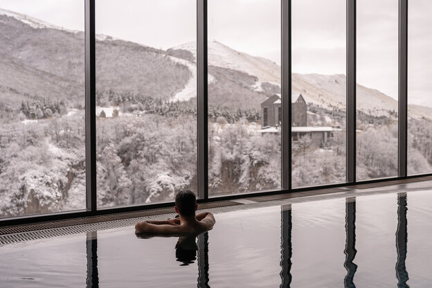 Ein Männerrücken ist im Schwimmbecken zu sehen, der Mann schaut auf die Berge durch riesige Glasfenster
