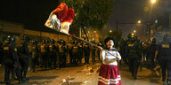 Ein Frau in indigener Kleidung schwenkt steht vor einer Reihe von Polizisten die peruanische Flagge
