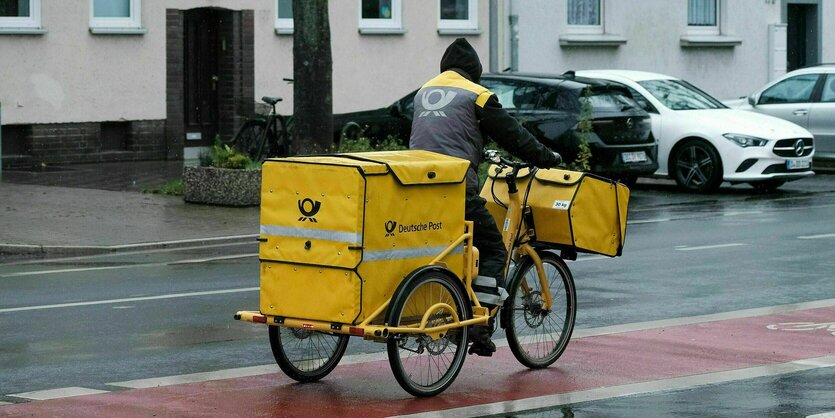 Briefzusteller radelt mit seinem Lastenfahrrad über eine regennasse Straße