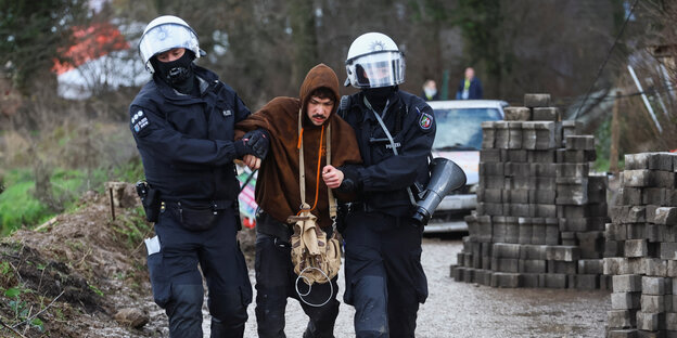 Zwei Polizisten mit Polizeihelmen führen einen jungen Mann mit braunem Kapuzenpullover ab.