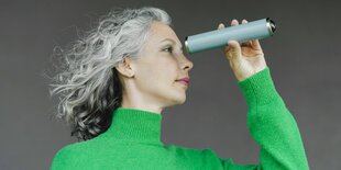 ein Frau mit längeren grauen Haaren trägt einen grünen Pullover und schaut durch Kaleidoskop