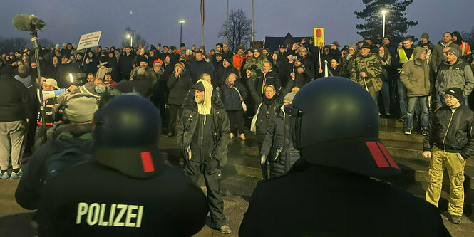 Polizisten mit m Rücken zur Kamera, dahinter Demonstrant:innen mit Schildern