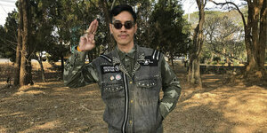 Der Angeklagte Mongkhon Thirakot steht mit dunkler Sonnenbrille und Jeansjacke vor Bäumen und macht das "Drei-Finger" Zeichen