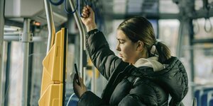 Eine junge Frau kauft sich mit ihrem Smartphone ein Ticket im Bus, während sie sich an einer Schlauf festhält