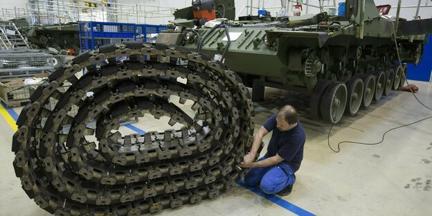 Ein Mann hat in einem Werk eine aufgerollte Panzerkette vor sich liegen und arbeitet daran, im Hintergrund steht ein Leopard-II-Panzer