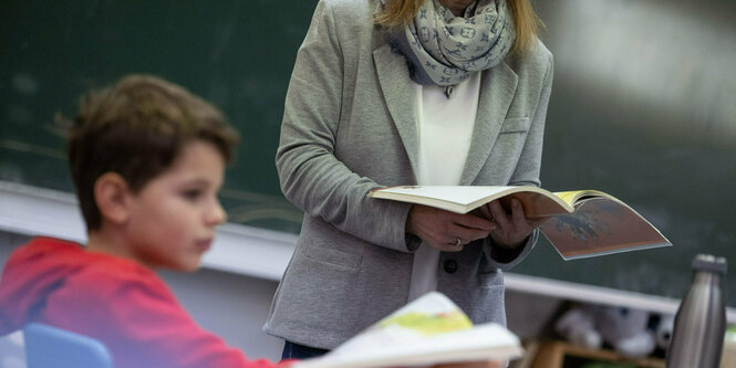 Eine Lehrerin mit Fibel steht neben eiem schüler, der auch eine aufgeschlagene Fibel auf dem Tisch hat