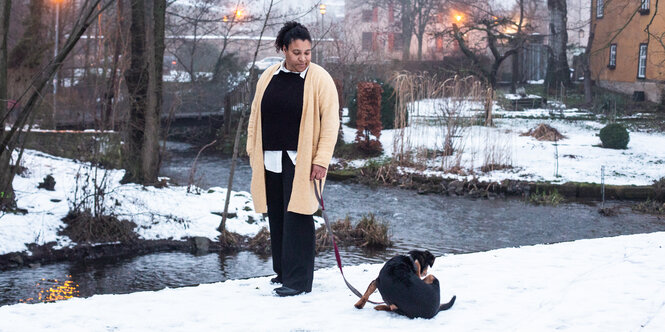 Eine Frau in gelber Jacke steht im Schnee und schaut auf ihren Hund