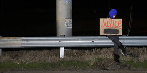 Eine Frau mit blauem Kopftuch sitzt nachts auf einer Leitplanke und hält ein Schild mit der Aufschrift „Justice“. Die Szene ist mit Scheinwerfern erleuchtet