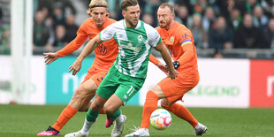 Zwei Fußballer in orangen Trikots laufen einem Fußballer in grünem Trikot hinterher, der den Ball spielt