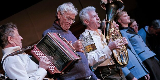 Gerhard Polt mit Bierglas steht zwischen zwei Musikern mit Akkordeon und einem Blasinstrument