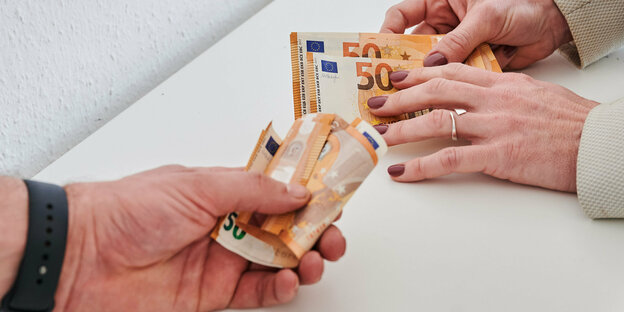 Eine Hand mit lackierten Nägeln und eine Hand mit schwerer Armbanduhr halten Geldscheine - letztere mehr als erstere.