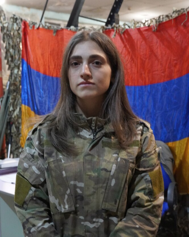 Eine junge Frau mit offenen Haaren im Tarnfarbenanzug vor der Flagge Armeniens