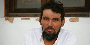Ein Mann mit sonnengebräuntem Gesicht sitzt vor einem Haus